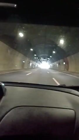 The video captures the interior view from a vehicle traveling through a well-lit tunnel. The tunnel has a circular or arched ceiling with numerous lights spaced evenly along its length. The walls appear to be made of concrete or a similar material and show some wear and tear. White lane markers are visible on the road, indicating multiple lanes of traffic. The car is moving forward, and the end of the tunnel, which is brightly illuminated, is visible in the distance, suggesting the driver is approaching the exit. There are no other vehicles or people clearly visible within the tunnel. The dashboard of the car is partially visible in the foreground, indicating the perspective is from the driver's seat.