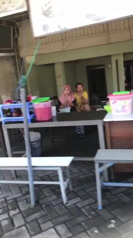 Two women are seated at a table in front of a building. The woman on the left is wearing a pink headscarf and a light-colored top. She is looking down at the table. The woman on the right is wearing a yellow top and is looking towards the left. There is a table with various items on it, including a pink bucket with a green lid, a clear container with a pink liquid, and some white cups. In the foreground, there are two metal tables with white tops. The ground is paved with bricks. The scene appears to be an outdoor food stall or eatery.