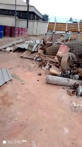 The video shows a cluttered outdoor area with a dirt ground. Various discarded items are scattered throughout the space, including pieces of metal, tires, pipes, wood, and wire mesh. There are also a few barrels visible in the background. The area is enclosed by a fence, and the overall impression is one of disarray and abandonment. There are no people visible in the video. The camera seems to be handheld and moves slightly, providing different views of the debris.