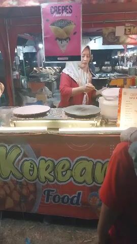 A woman wearing a red shirt and a white hijab is preparing crepes at a food stall. She is standing behind a counter that has the words "Korean Food" written on it in large, colorful letters. There are two crepe makers on the counter, and the woman is using a ladle to pour batter onto one of them. A sign above her reads "Crepes Food Menu" with an image of a chocolate-covered crepe. In the background, there are other food stalls and people. The scene appears to be at a night market or outdoor food festival.