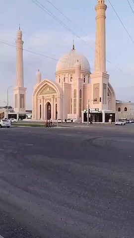 The video shows a mosque from the street. The mosque is a light tan color with a large dome and two minarets. There are people walking around the mosque and cars driving on the street. There is a small market next to the mosque.