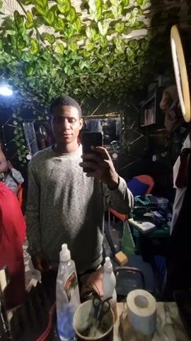 A man takes a selfie in a mirror in what appears to be a salon. He is wearing a heather grey long-sleeved shirt. The salon features faux ivy on the ceiling, creating a jungle-like atmosphere. There is a ring light and other salon supplies, such as bottles, scissors, and paper towels, around him.  Another person is visible in the background wearing a red garment.