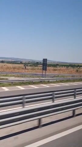 A view from a moving vehicle shows a highway with metal guardrails. Beyond the guardrails, a dry, grassy field stretches to a distant line of trees and hills under a clear blue sky. A road sign is visible in the distance. A truck and a blue car pass by in the opposite lanes. Several tall trees are present on the left side of the frame.
