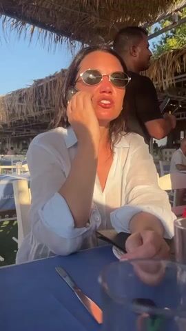 A woman with dark hair and sunglasses is sitting at a table under a thatched roof, talking on a cell phone. She is wearing a white semi-sheer shirt. There is a knife and a glass on the blue tablecloth in front of her. In the background, other people are seated at tables, and a man wearing a black shirt is visible to her right. The lighting suggests it is late afternoon or early evening.