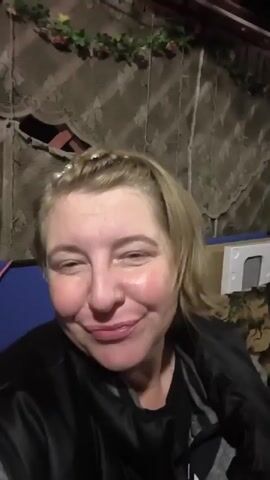 The video shows a woman with blonde hair and a pearl headband. She is wearing a black jacket over a grey and black striped shirt. She is making faces at the camera, including sticking out her tongue. The background appears to be a dimly lit indoor space with draped fabric and some objects that are difficult to identify. The woman is the sole focus of the video.
