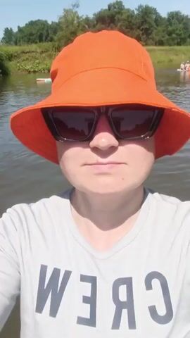 A person wearing an orange bucket hat and sunglasses is looking at the camera. They are on a body of water, likely a river, with other people on paddleboards in the background. The water is brown and there are trees and green foliage on the banks of the river. The person in the foreground is wearing a grey t-shirt with "CREW" written on it in black letters.