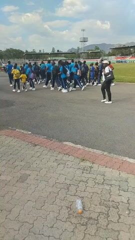 The video depicts a large group of people, mostly adults, walking together in what appears to be the parking lot of an outdoor stadium. They are dressed in a variety of clothing, with many wearing blue or yellow shirts and dark pants. Some individuals carry backpacks. A person in a white cap, black top, and black pants walks perpendicular to the main group. The ground is paved with light grey bricks outlined with a red trim. In the foreground, a clear plastic water bottle lies on the ground. Beyond the parking lot is a green sports field with spectators in the stands and a tall antenna tower. The sky is partly cloudy.