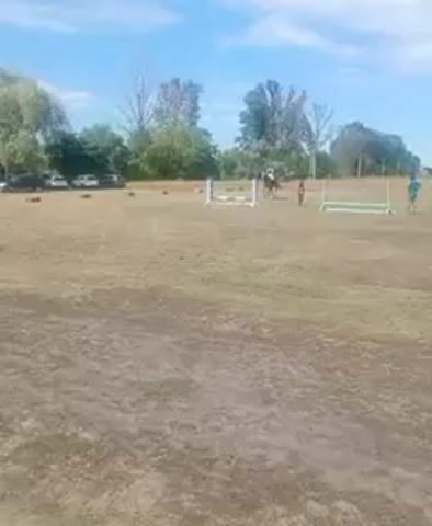 The video shows an outdoor equestrian event. In the foreground, a dry, grassy field with patches of dirt is visible. In the mid-ground, several horses and riders are present, along with equestrian jumps. One horse and rider are in the process of jumping over a white obstacle. Another rider on a brown horse is approaching a jump. In the background, more horses and riders are visible, as well as parked cars and trees. The sky is blue with some scattered clouds. The overall scene suggests a competition or training session for horse jumping.