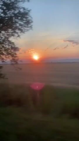 The video shows a sunrise over a rural landscape. The sun is low on the horizon, casting a warm orange glow across the sky. The sky is a mix of blue and pink, with a few scattered clouds. In the foreground, there are trees and fields, blurred due to the motion of the camera. The overall impression is one of peace and tranquility.