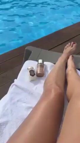 A person is lounging on a white towel on a brown deck next to a pool. Two bottles are also on the towel. The person has tan legs and red nail polish on their toes. The pool is full of people swimming and relaxing. A lush green mountain sits in the background under a cloudy sky.