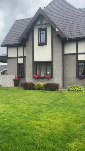 A drone shot moves slowly over a well-manicured green lawn towards a two-story house with a brown roof and beige and grey siding. The house has several windows with flower boxes filled with pink and red flowers. There are also large planters with red flowers on the ground. To the left of the house, there is a covered porch area. In the background, other houses and trees are visible under a cloudy sky.