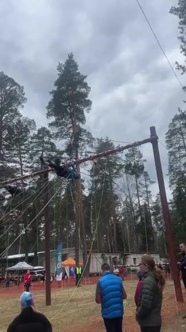 The video shows a low-resolution view of an outdoor obstacle course with several participants. Two individuals are traversing an elevated section involving ropes and beams, suspended upside down and using their hands to move across. The people appear to be wearing helmets and harnesses. At ground level, a small crowd of spectators watches, including people in casual attire like jackets, vests, and hats. The scene is set in a wooded area with tall pine trees and a grassy field. In the background, there are tents and parked vehicles. The sky is overcast, suggesting a cloudy day.
