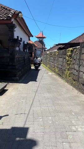 The video shows a narrow paved alleyway in a tropical location with a clear blue sky. Three people on motorbikes are driving away from the camera. The alley is bordered by stone walls and traditional Balinese structures with red tile roofs. One wall has moss growing on it. There are power lines overhead. The scene is well-lit.