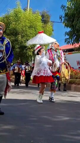 A parade is taking place outdoors on a sunny day. In the foreground, two individuals in traditional costumes are dancing. The person on the left is wearing a blue jacket, white pants, and a hat, with a mask covering their face. The person on the right, a child, is wearing a white and red dress, a hat, and holding a white umbrella. Behind them, a group of musicians is walking, playing instruments including accordions and drums. The musicians and other parade participants are also wearing colorful costumes. The background shows trees, buildings, and a clear blue sky.
