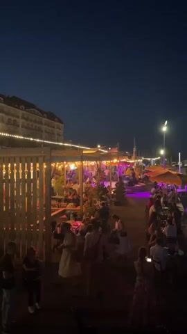 The video shows a lively outdoor scene at night, likely a waterfront restaurant or bar. A crowd of people is seated at tables, illuminated by string lights and colorful uplighting, creating a festive atmosphere. The architecture in the background appears to be a building with multiple floors and balconies. Adjacent to the dining area, a harbor is visible with several boats docked. The water reflects the ambient light, and a few distant lights suggest the presence of a town or city. The overall scene depicts a social gathering in a pleasant evening setting.