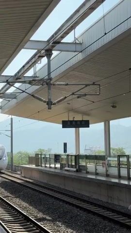 A high-speed train with blue and white markings passes by a train station platform. The station has a covered roof supported by pillars, a safety barrier along the platform edge, and tracks in the foreground. Beyond the station, there are buildings and a forested hillside under a hazy sky.