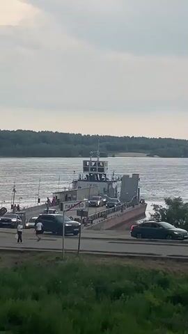 A ferry boat is docked by the shore, carrying several cars and passengers. A few cars are waiting on the road next to the ferry, and some people are walking around. The ferry has a white cabin structure on top and a ramp that is slightly raised. The water is calm and wide, with a lush green forest on the opposite bank. The sky is overcast with clouds.