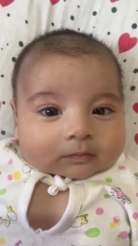 The video is a close-up, head-on shot of a baby's face. The baby has dark hair, dark eyes, and a light complexion. The baby is wearing a white onesie with a pattern of hearts and circles. The background is a white surface with a pattern of red hearts and black dots. The baby's facial expression changes slightly throughout the video, shifting from neutral to a slight frown and then back to neutral. The lighting is bright and even.