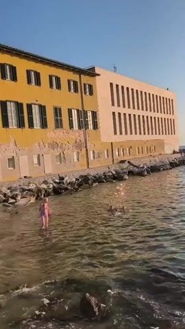 A drone shot pans over a body of water with a young child in a pink swimsuit standing in the shallow water on the left, holding a ball. Two other individuals, one possibly an adult and another child, are further out in the water, swimming. A rocky shoreline runs along the water's edge, leading to a yellow building with arched windows and shutters, followed by a pale peach-colored building with a grid of rectangular windows. The sky is clear and blue, and the water appears calm with gentle ripples. The scene suggests a sunny day at a coastal location.