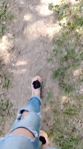 A first-person point-of-view shot shows a person walking on a rocky path. The person is wearing light blue ripped jeans and beige sandals with black pom-poms on the toes. Green grass and scattered leaves are visible on either side of the path. The person is walking forward, their feet moving one after the other.