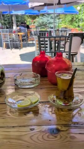 The video pans across a charming outdoor seating area of a cafe in Şəki, Azerbaijan. In the foreground, a wooden table displays several items, including a white teapot with a floral pattern, an ashtray, a small glass dish with lemon slices, and a tall glass filled with amber-colored liquid, likely tea, with a cinnamon stick or similar stirrer. Two bright red pomegranates are also prominent on the table.

The camera then moves to reveal the wider setting. Several tables and chairs are arranged on a paved courtyard, shaded by large blue and black umbrellas. In the background, people are seated at tables, engaged in conversation or enjoying their time at the cafe. Lush green trees provide further shade and a pleasant ambiance. String lights are visible overhead, suggesting the cafe is also operational in the evening. The architecture in the background hints at a traditional Azerbaijani style.

The overall mood is relaxed and inviting, characteristic of a pleasant afternoon spent at an outdoor cafe. The sunlight filtering through the trees indicates it is daytime, likely late morning or early afternoon, with good weather. No visible text is present in the video.