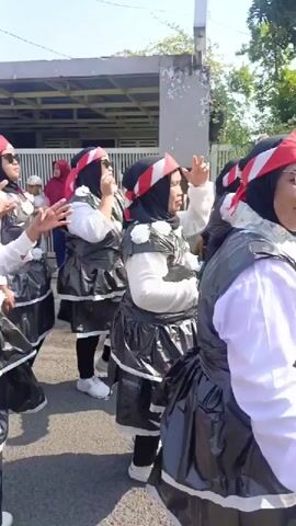 This is a close-up, medium shot video taken from a low angle. It shows a group of people, mostly women, dressed in costumes. They are wearing white shirts, black vests with white trim, black skirts made of shiny material, and red, white, and black hats. Some of the women are wearing sunglasses. They are walking in a procession on an asphalt street. In the background, there are houses and a fence. The sky is clear and blue. The people in the video appear to be smiling and enjoying themselves. The overall atmosphere is festive.