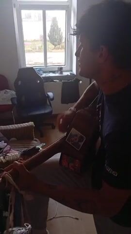 A young man with dark, wavy hair sits indoors playing an acoustic guitar. He is wearing a dark t-shirt and light-colored pants. The guitar has stickers on it. He is looking to his right and appears to be singing or speaking. The room is lit by natural light coming from a window. In the background, there is a black gaming chair, a couch with striped cushions, a bed, and clothes scattered around. The overall environment appears to be a bedroom or living space.