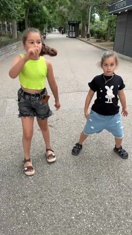 Two young girls are dancing on a paved walkway in a park. The girl on the left is wearing a neon green tank top and denim shorts. The girl on the right is wearing a black t-shirt with a white bunny graphic and light blue denim shorts. Both girls are wearing sandals. The background shows trees, a sidewalk, and a building. The girls are facing forward and appear to be in motion, with their arms and legs extended as if performing a choreographed dance. The lighting suggests it is daytime.