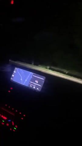 The video captures a first-person perspective from inside a moving vehicle at night. A digital display, likely a car's infotainment screen, is visible on the lower left of the frame, showing an interface with text and symbols. The car's headlights illuminate a dark asphalt road ahead, which has a faded white center line. Trees and indistinct dark shapes line the sides of the road. The surrounding environment is predominantly dark. Audio features muffled speech and music playing within the vehicle.