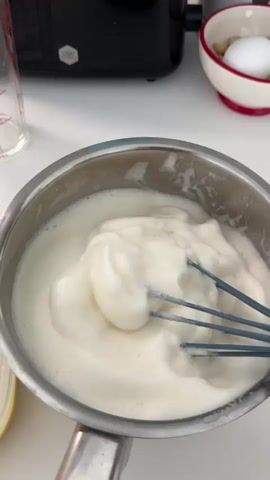 A close-up shot shows a whisk with a light-colored foam being lifted from a silver saucepan. The saucepan contains more of the same frothy substance, which appears to be a mixture of liquid and air. The whisk itself has a blue handle and metal wires. In the background, a black appliance is visible, along with a white bowl containing a white egg and some nuts. A glass measuring cup is also partially visible. The overall setting suggests a kitchen environment where food preparation is taking place.