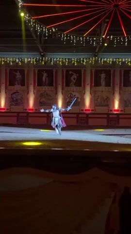 A man dressed as a gladiator with a sword and a red cape is performing in an arena. He is surrounded by sand and columns, with murals on the wall behind him. The lighting is dramatic, with spotlights illuminating the performer. Another man in a blue sash is also present in the arena. The video appears to capture a reenactment or performance of a gladiatorial combat.