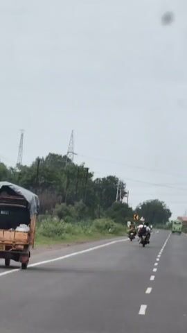 A first-person perspective video shows a paved road with a white dashed line in the center. There are several motorcycles with riders on the road, moving away from the camera. The road is lined with green vegetation on the left and right. Tall power lines and towers are visible in the background against a cloudy sky. On the right side of the road, a green bus is also moving away from the camera. A truck with a covered bed is partially visible on the left side of the road, also moving away from the camera.