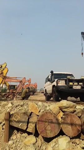 The video shows a construction site on a clear day with a pale blue sky. In the foreground, there are several large, cut logs piled up, along with rocky debris. A person wearing a green shirt and dark pants is crouched down, appearing to be working on something near the logs. Behind them, a white SUV with off-road modifications, including a bull bar and extra lights, is parked on a dirt road. Several large orange excavators are visible further in the background, suggesting ongoing construction or demolition work. A crane can be seen in the far distance. The overall impression is one of an active industrial or construction environment.