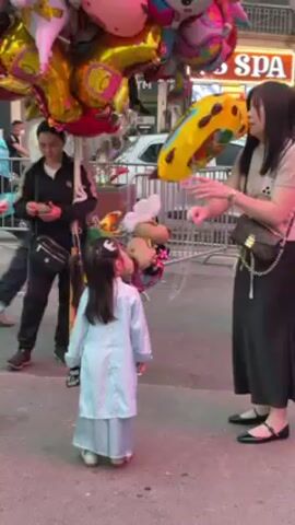 The video shows a young girl wearing a light blue traditional Vietnamese outfit and standing in front of an adult woman who appears to be purchasing a balloon from a balloon vendor. The balloon vendor holds a large bunch of colorful foil balloons in various shapes, including a giraffe and a unicorn. The woman buying the balloon is wearing a light-colored t-shirt, a long black skirt, and black flats, and she is carrying a purse with a chain strap. She appears to be handing money to a man who is likely the balloon vendor, as he is partially visible to the left and seems to be receiving the payment. In the background, there are other people, including a woman in a red top and another person holding a small cup or snack. The scene seems to be taking place on a street, with a spa sign visible in the background and a car parked nearby. The general atmosphere appears to be a festive or public gathering.