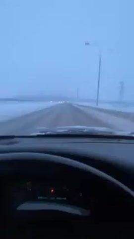 The video shows a driver's perspective from inside a car driving on a snowy road. The sky is overcast, giving a bluish-gray cast to the scene. The road is straight and stretches into the distance, with snow-covered ground on either side. Streetlights are visible along the road. The car's dashboard is visible in the foreground, and the windshield wipers are active, indicating snow or ice on the windshield.