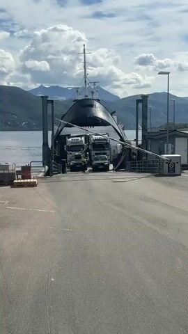 Two large trucks are positioned on a ferry ramp. A person in a green shirt and a hard hat is standing between the two trucks. The ferry is dark and appears to be a roll-on/roll-off ferry. Mountains and a body of water are visible in the background. The sky is cloudy. A gate is partially lowered on the right side of the ramp. The ground is paved.