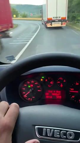 The video shows a first-person view from inside a truck. The driver's hands are on the steering wheel, which has a dashboard with red illuminated gauges and a digital display. The truck is moving on a road with a truck in front of it. The road curves to the left, and there are trees and hills on the side. The side mirror reflects other vehicles, including trucks and cars, in a line on the road ahead, suggesting a traffic jam. The weather appears to be overcast, and the time of day seems to be late afternoon or early evening.