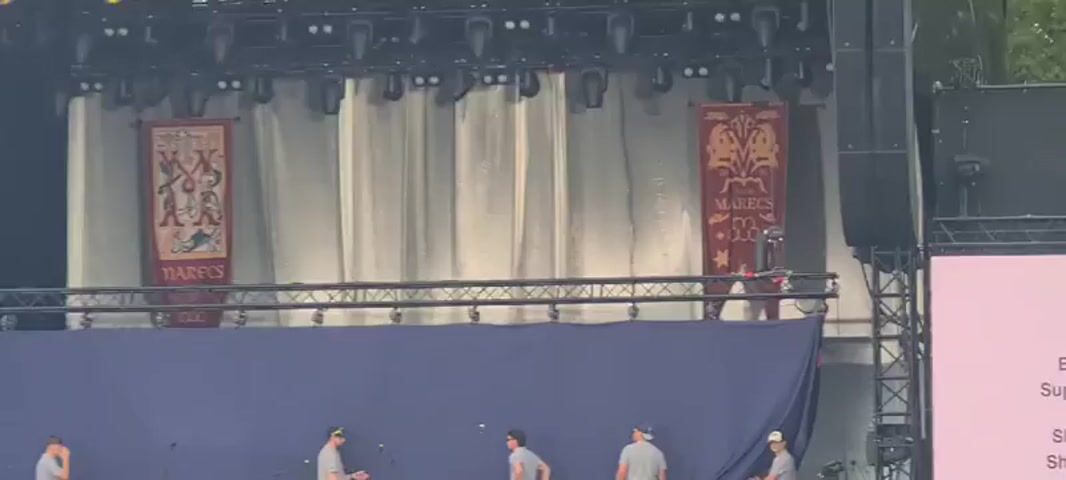 The video shows a group of four men on a stage setting up a large blue backdrop. Two banners hang behind the backdrop, one on the left with figures and text that appears to say "MARECS LOV" and another on the right with a similar design and text that reads "MARECS LO." There are also drums and other stage equipment visible. The men are wearing casual clothing, with three of them wearing gray t-shirts and one wearing a blue t-shirt. One man on the left is near the drums, another in the center appears to be looking at the backdrop, and two men on the right are holding and adjusting the backdrop. In the background, there is lighting equipment and a sound system. A drone is also visible hovering near the right banner.
