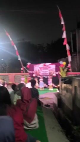 A nighttime outdoor performance is underway, with a stage set up under a dark sky. Indonesian flags, red and white, are prominently displayed on tall poles, flanking the stage area. The stage itself has a banner that reads "TIRAKATAN HUT RI KE-78" which translates to "Celebration of the 78th Anniversary of the Republic of Indonesia," indicating a national holiday event. Several young children, dressed in costumes, are performing on stage. The audience, seated on the ground, is visible in the foreground, with some individuals wearing red. Lighting on stage illuminates the performers. The environment appears to be a community gathering space.