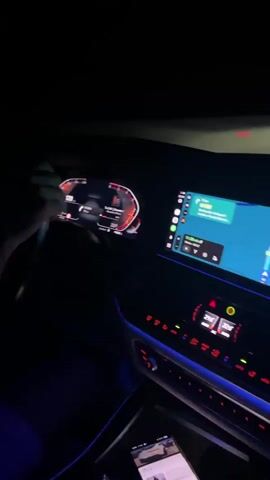 The video shows the interior of a car at night. The dashboard and infotainment screen are illuminated with ambient lighting and displays. A person is driving, their hands on the steering wheel. Another person is in the passenger seat, wearing glasses and a white t-shirt. The ceiling of the car has a star-like illuminated pattern. The infotainment screen displays a navigation map.