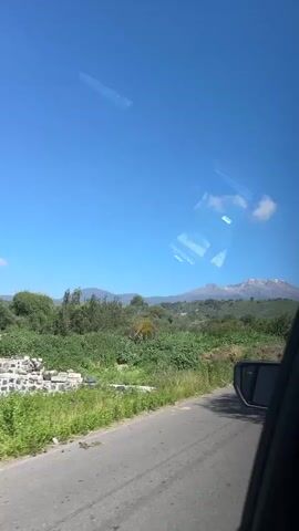 A video captured from inside a vehicle shows a view of a landscape through the windshield and side window. The sky is clear and blue. In the distance, a snow-capped mountain range is visible. In the foreground, there are fields of green crops, possibly corn, and various trees and vegetation. A side mirror of the vehicle is also visible on the right side of the frame. A reflection of the interior of the vehicle, including what appears to be part of a dashboard or seat, is visible on the windshield. The video appears to be filmed while the vehicle is in motion, suggesting a roadside or highway view.