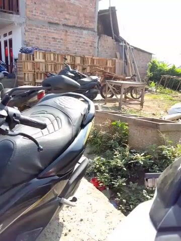 The video shows an outdoor scene with several motorcycles parked in the foreground and middle ground. In the background, there is a brick building with a wooden balcony, and next to it, a smaller building with a corrugated metal roof. Several wooden crates are stacked near the buildings. There are also a few people visible, some of whom appear to be interacting with each other or with the motorcycles. The environment appears to be somewhat rural or semi-urban, with some vegetation and uneven ground. The lighting suggests it is daytime, possibly late morning or early afternoon.