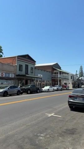 A vertical video pans across a street lined with old-fashioned buildings. The sky is clear and blue. Cars are parked along the street, and some buildings have signs for businesses such as "Ione Auto Parts" and "Donuts." The overall impression is of a quiet, small-town street on a sunny day.