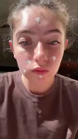A fair-skinned woman wearing a brown t-shirt is seen applying a white substance to her face. The substance is applied in small dots on her forehead, cheeks, and chin. She uses her right hand to rub the substance into her forehead in an upward motion. Her nails are decorated with colorful designs. She has a nose piercing and braces are visible on her teeth. The background is out of focus and appears to be indoors. A loud sound is audible.