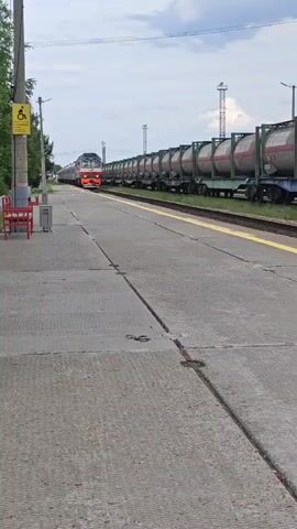 The video shows a train with a locomotive and multiple tank cars stopped at a train station. The platform is concrete with yellow lines. There are benches, a trash can, and a sign with a wheelchair symbol visible. The sky is cloudy. The train is red and white, and the tank cars are green with red markings. The train appears to be stationary at the station.