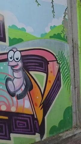 The video shows a graffiti mural on a wall. The mural depicts a cartoonish worm with big eyes and a smiling face. The worm is positioned in the center of the frame, with a vibrant and colorful background. The background features abstract shapes, swirling patterns, and various shades of pink, orange, green, and purple. The mural also includes elements of a landscape with rolling hills and a sky, suggesting an outdoor setting. The camera pans across the mural, highlighting different sections and details of the artwork. The wall appears to be made of concrete, and a portion of the ground, covered with brick paving stones, is visible at the bottom of the frame. There are no people or animals present in the video, only the painted artwork.