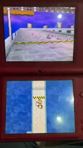Here is a description of the video:

The video begins with a close-up shot of a red Nintendo 3DS, displaying the game Mario Kart. The upper screen shows the racing scene, while the lower screen displays the track layout. The camera then pans up to reveal the surrounding environment: a large, white, tent-like structure. Inside, there are rows of green cots with green duffel bags and other personal belongings. Several people in military uniform are present, some standing and conversing, while others appear to be resting. A barefoot individual is visible in the foreground. Through the tent's opening, there is a glimpse of military vehicles. The overall setting suggests a military or refugee camp.