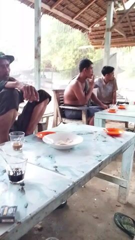 The video shows an outdoor, partially covered space with four people sitting at two tables. Two of the people are shirtless, one is wearing a t-shirt, and the fourth is in a long-sleeved shirt. They appear to be drinking coffee or tea. There are motorcycles parked nearby. It is raining. The space is covered with a simple, weathered roof and netting for shade. There are cigarettes and a lighter on the tables.