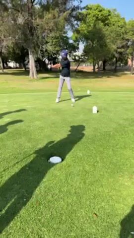 On a sunny day, a person wearing a long-sleeve shirt, light gray pants, white shoes, and a baseball cap swings a golf club on a green golf course. The person stands between three white tee markers, the middle of which has a golf ball on it. Trees are in the background. The person swings the club, and the video ends.