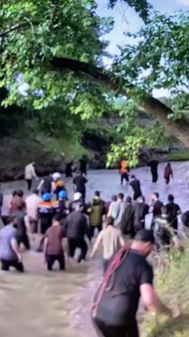 A group of people are wading through a shallow, muddy river. The riverbanks are lined with trees and vegetation. Some people are carrying bags and other belongings, suggesting they may be migrating or traveling. The water appears to be calm, and the people are moving in various directions. The scene is blurry, possibly due to the movement of the camera or the subjects.
